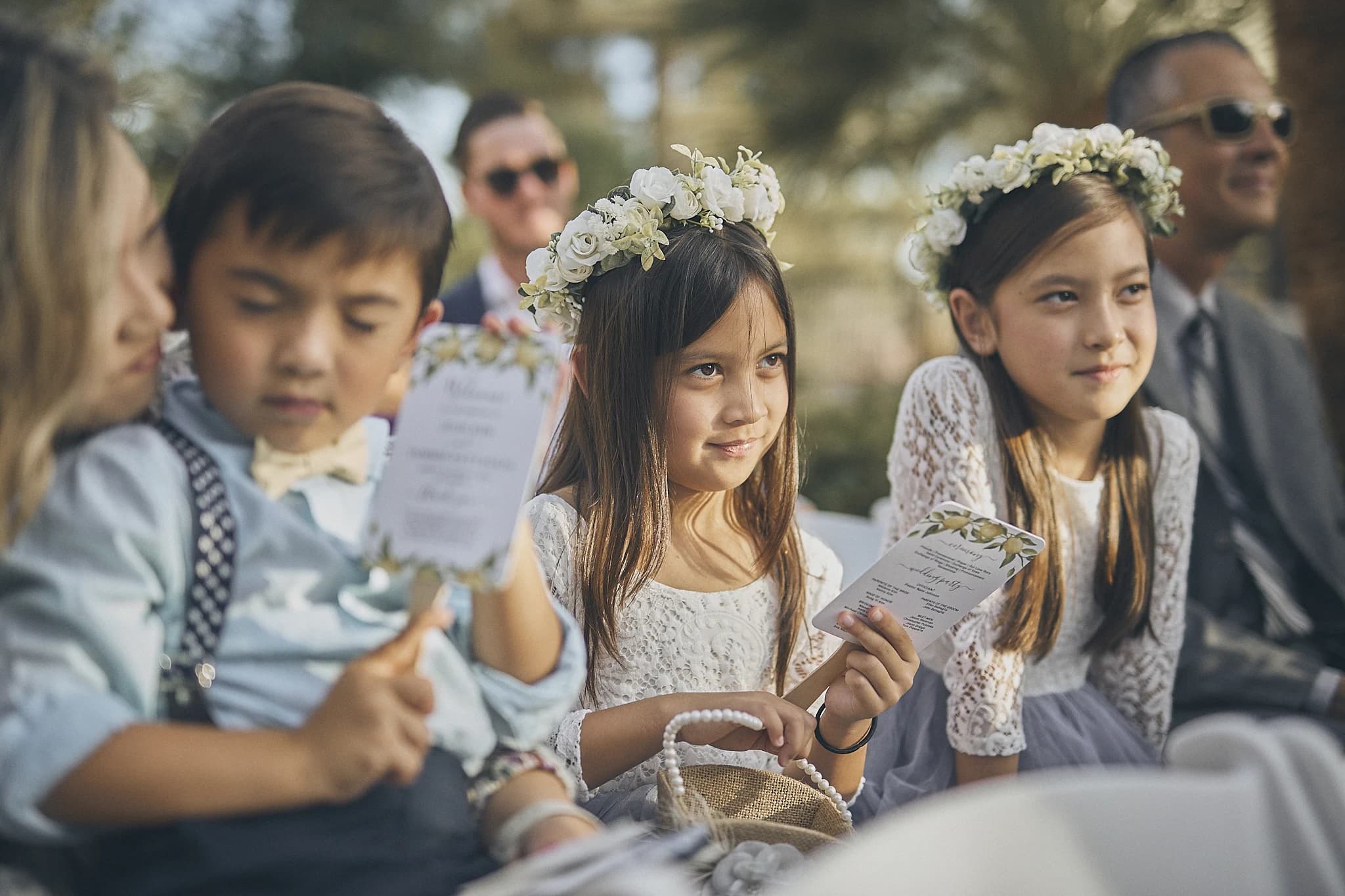 Children wearing floral crowns sit together during an outdoor ceremony