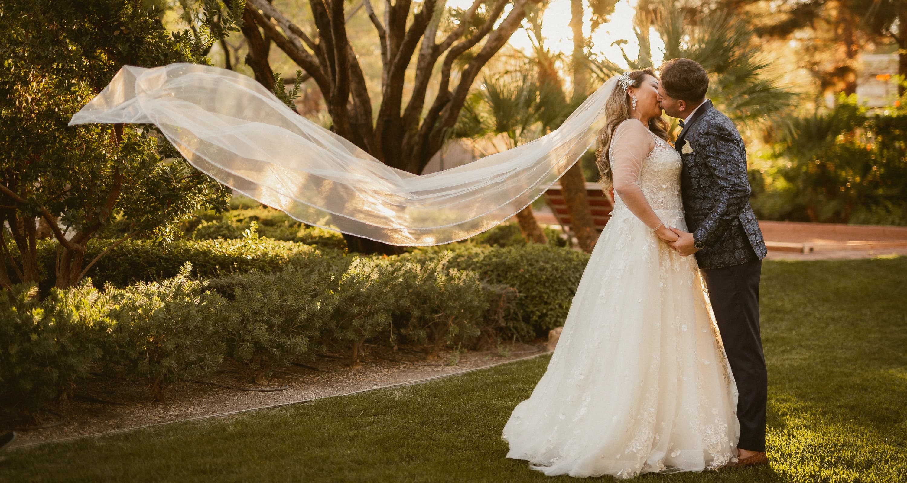 Newlyweds kiss in a sunlit garden as the bride's veil flows across the frame